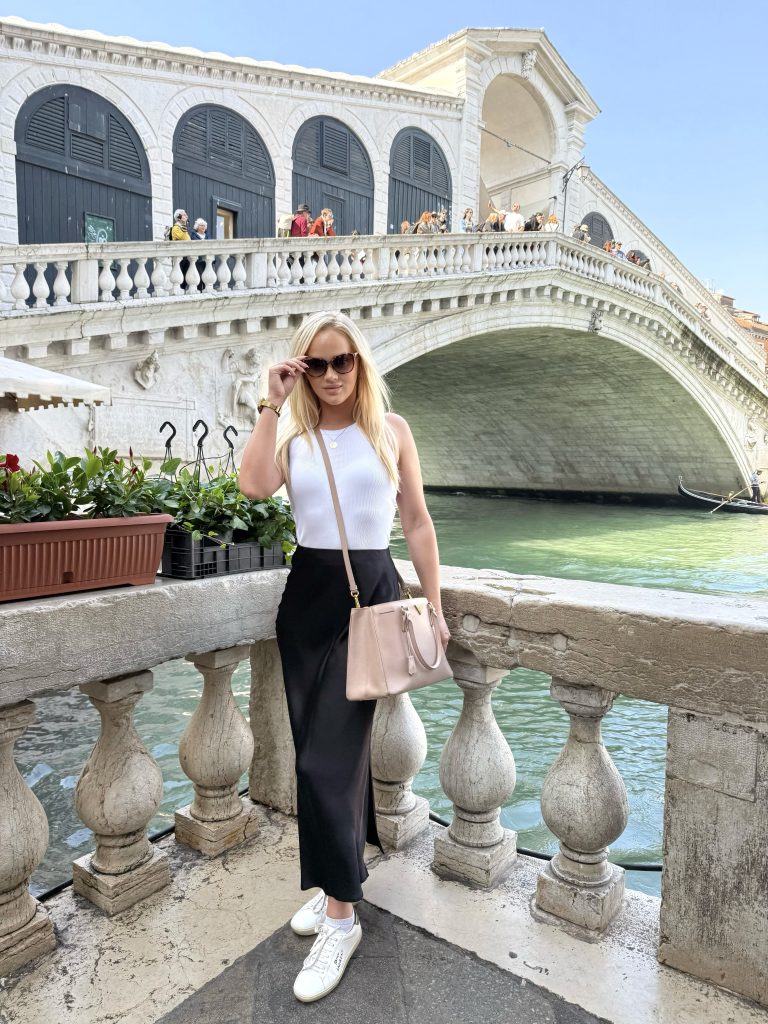 girl standing at Rialto bridge in Venice wearing outfit of black satin skirt, white racerback vest and white trainers by Saint Laurent