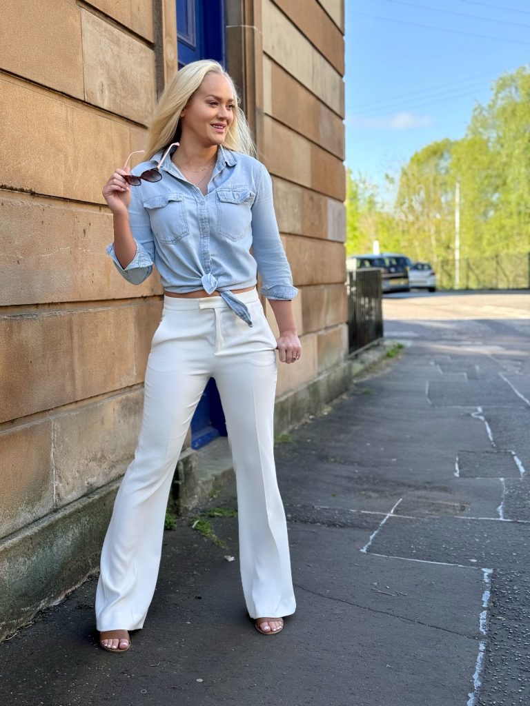 Girl's spring outfit is denim shirt and white trousers. She smiles as she removes sunglasses