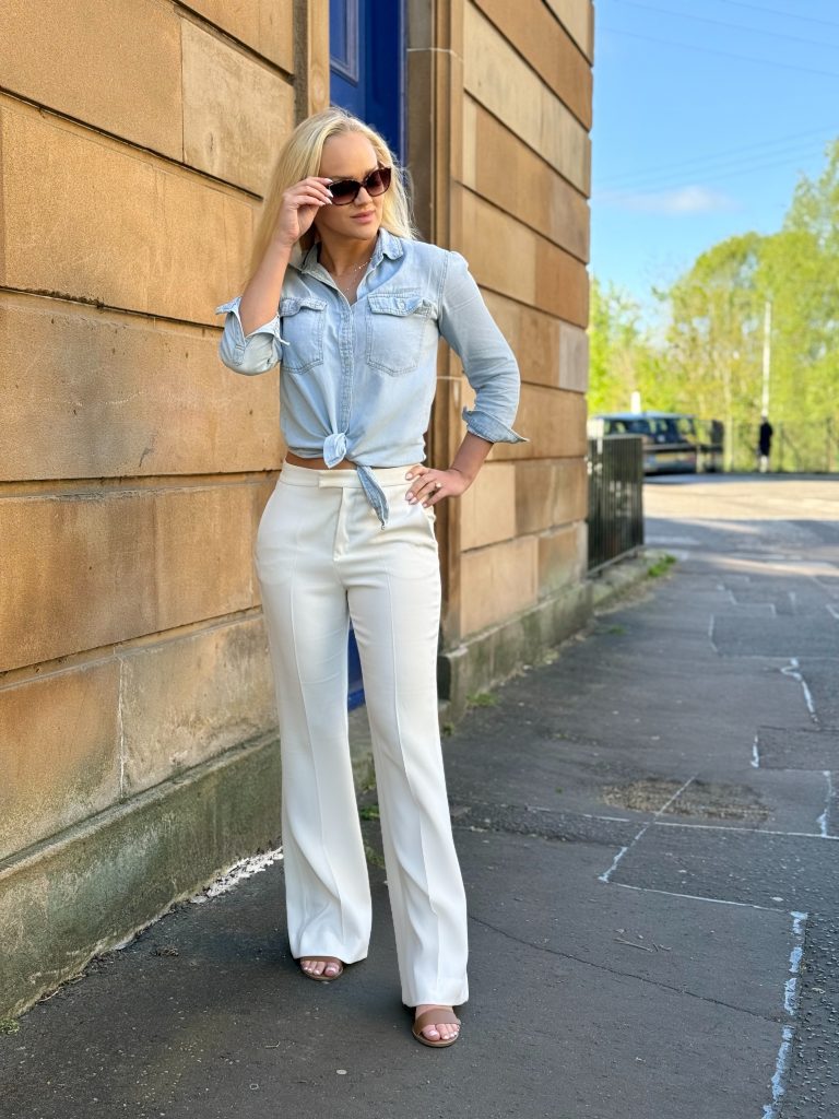 Girl on street wears relaxed denim shirt tied at waist with tailored white trousers