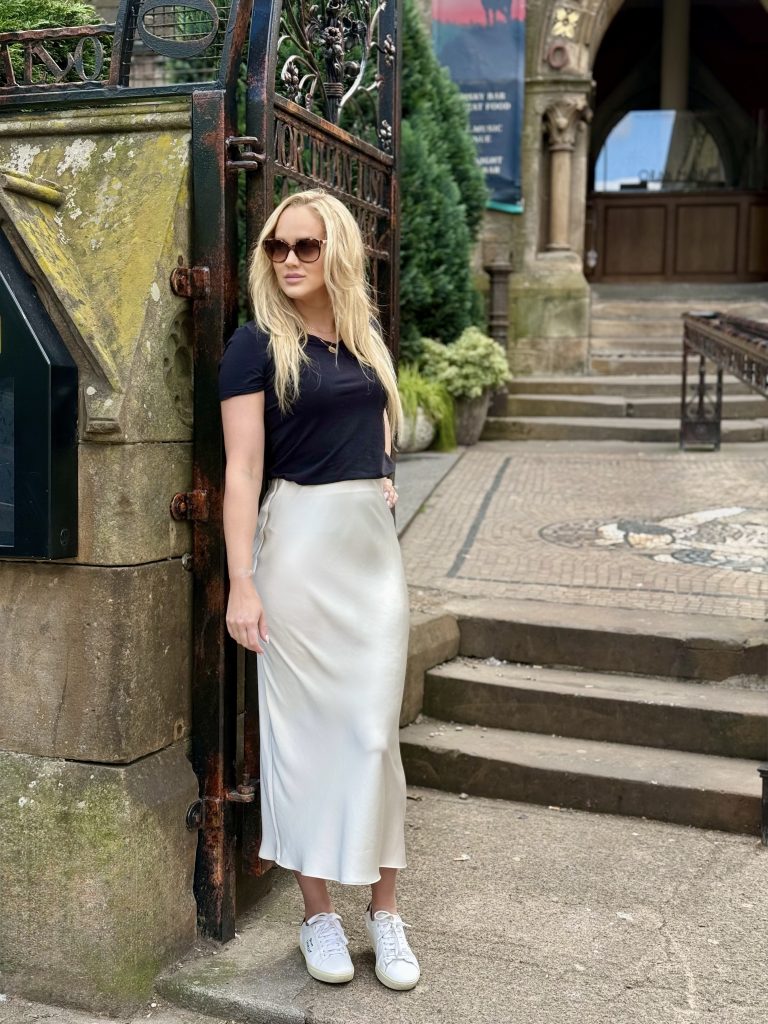 Girl wearing cream satin maxi skirt and black t-shirt with white trainers, leaning against a gate